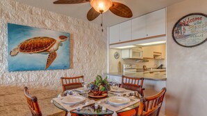 Zona pranzo con tavolo in vetro apparecchiato per quattro, circondata da sedie in vimini. Sul muro, un dipinto raffigura una tartaruga marina, e si legge un cartello di benvenuto per la casa sulla spiaggia.