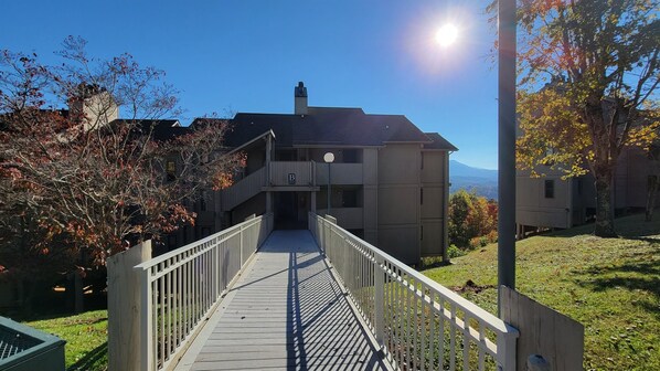 A wooden walkway leading to a building with a balcony, surrounded by trees and a clear sky.