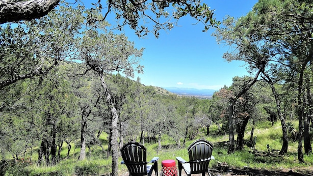 Eine natürliche Landschaft mit zwei Adirondack-Stühlen und einem roten Tisch, umgeben von Bäumen und einem entfernten Blick auf Berge.