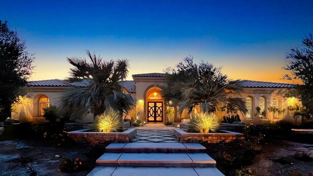 A well-lit entrance with a stone pathway leading to a gated door, surrounded by landscaped gardens and palm trees.