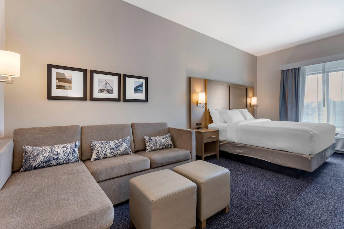 Hotelkamer met een slaapbank, voetenbankje, bed, nachtkastjes, wandlampen, ingekaderde foto's en een groot raam.