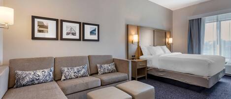 A hotel room with a sofa, ottoman, bed, nightstands, wall-mounted lamps, framed pictures, and a large window.