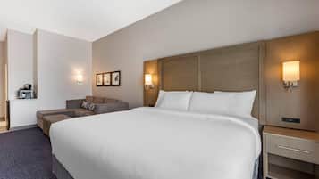 Hotellihuone, jossa on suuri sänky, sohva, työpöytä lampun kanssa ja seinälle kiinnitetty hylly, jonka päällä on kahvinkeitin.