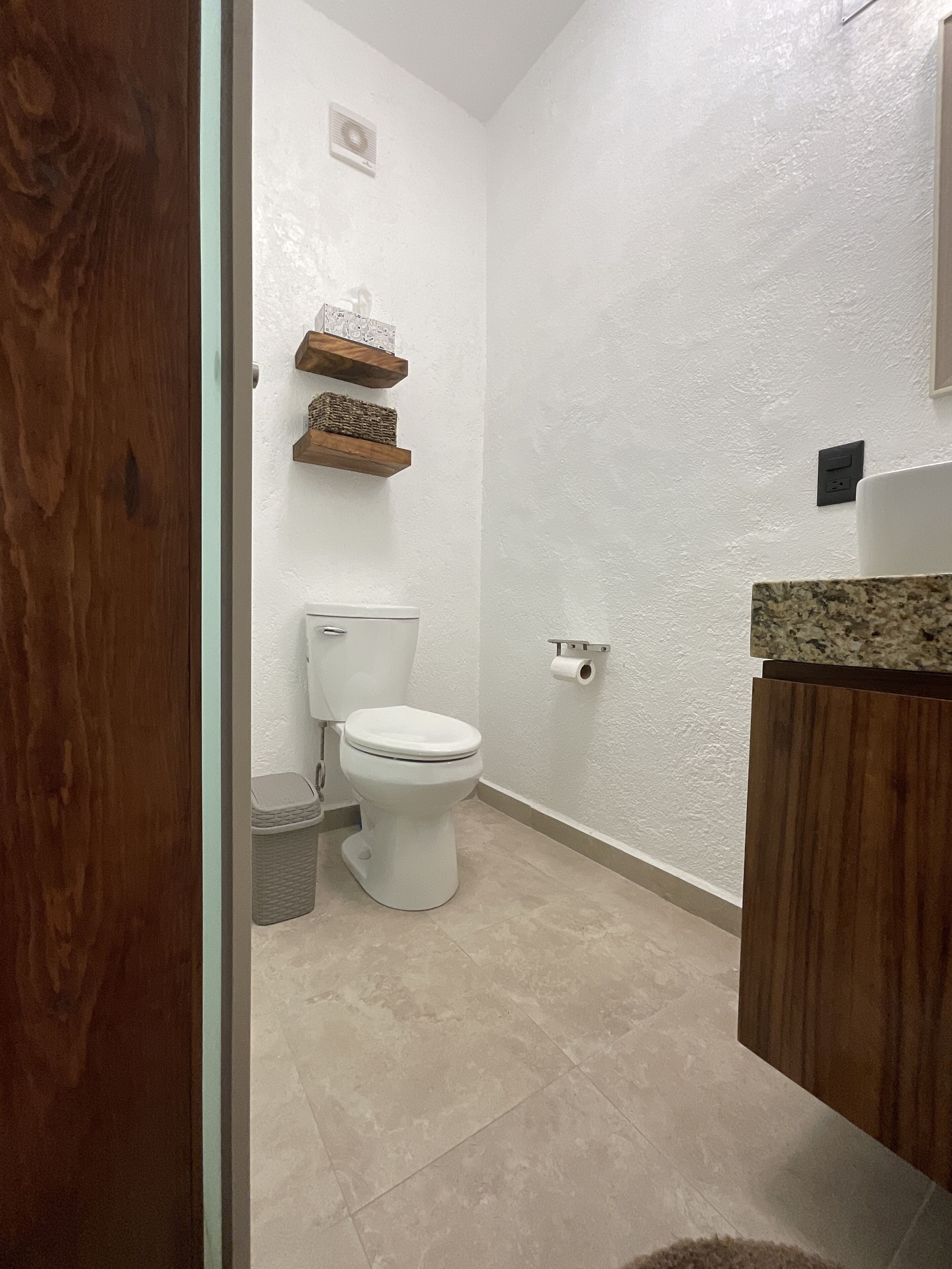 A bathroom with a toilet, a trash bin, a wooden shelf with decorative items, and a marble countertop.