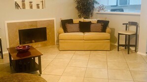Un salon avec un canapé beige, une table basse en bois, une cheminée, un ventilateur de plafond et un coin cuisine visible à travers une porte ouverte.