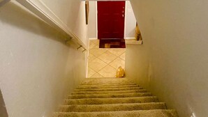 Un escalier recouvert de moquette, avec des murs blancs et une porte rouge en haut.