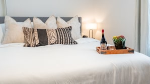 En pænt redt seng med mønstrede puder, et sengebord med en lampe, et bakke med en flaske og blomster, samt et natbord med en vase.