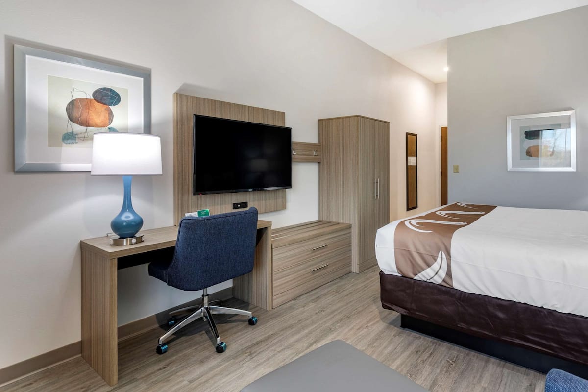 Ein Hotelzimmer mit Holzschreibtisch, einem Sessel, einem Flachbildfernseher, einem Bett mit braunem Kopfteil und einem gerahmten Bild an der Wand.