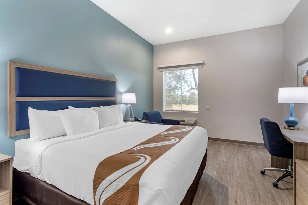 A hotel room with a large bed, a desk with a chair, a window, and a nightstand with a lamp.