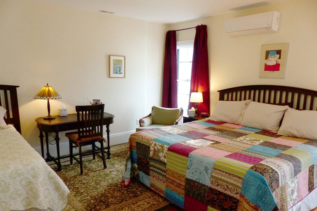 Une chambre à coucher avec un grand lit, un bureau avec une chaise, une table de chevet, une lampe et une fenêtre avec des rideaux.
