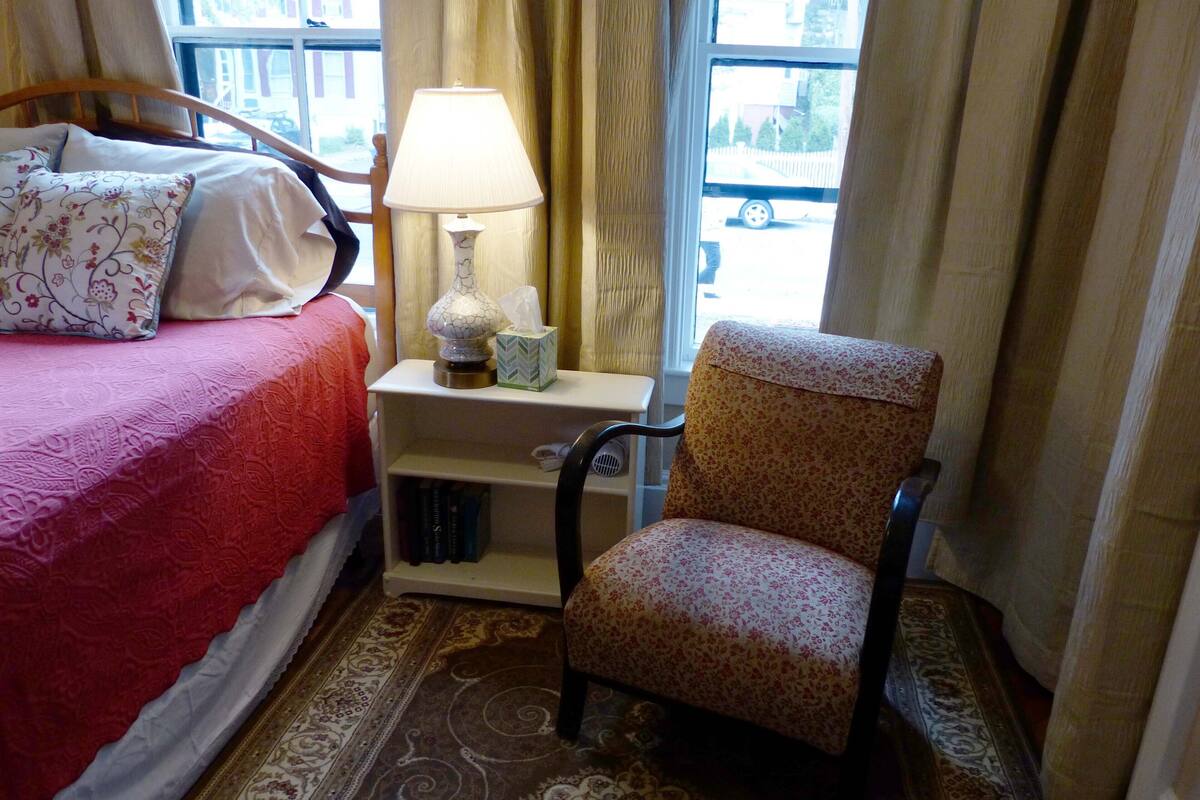 Une chambre d’hôtel avec un lit, une chaise, une table de chevet, une lampe et une fenêtre avec des rideaux.