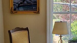 A room with a wooden chair with a cushion, a painting on the wall, a lamp on a table, and a window with a view of trees. - Davis Square Inn (Somerville)