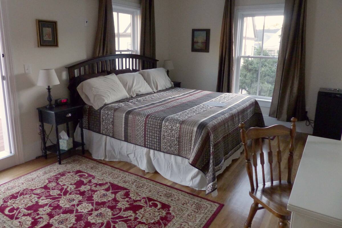 Une chambre à coucher avec un grand lit, deux tables de chevet, une chaise, un bureau et une fenêtre avec des rideaux.