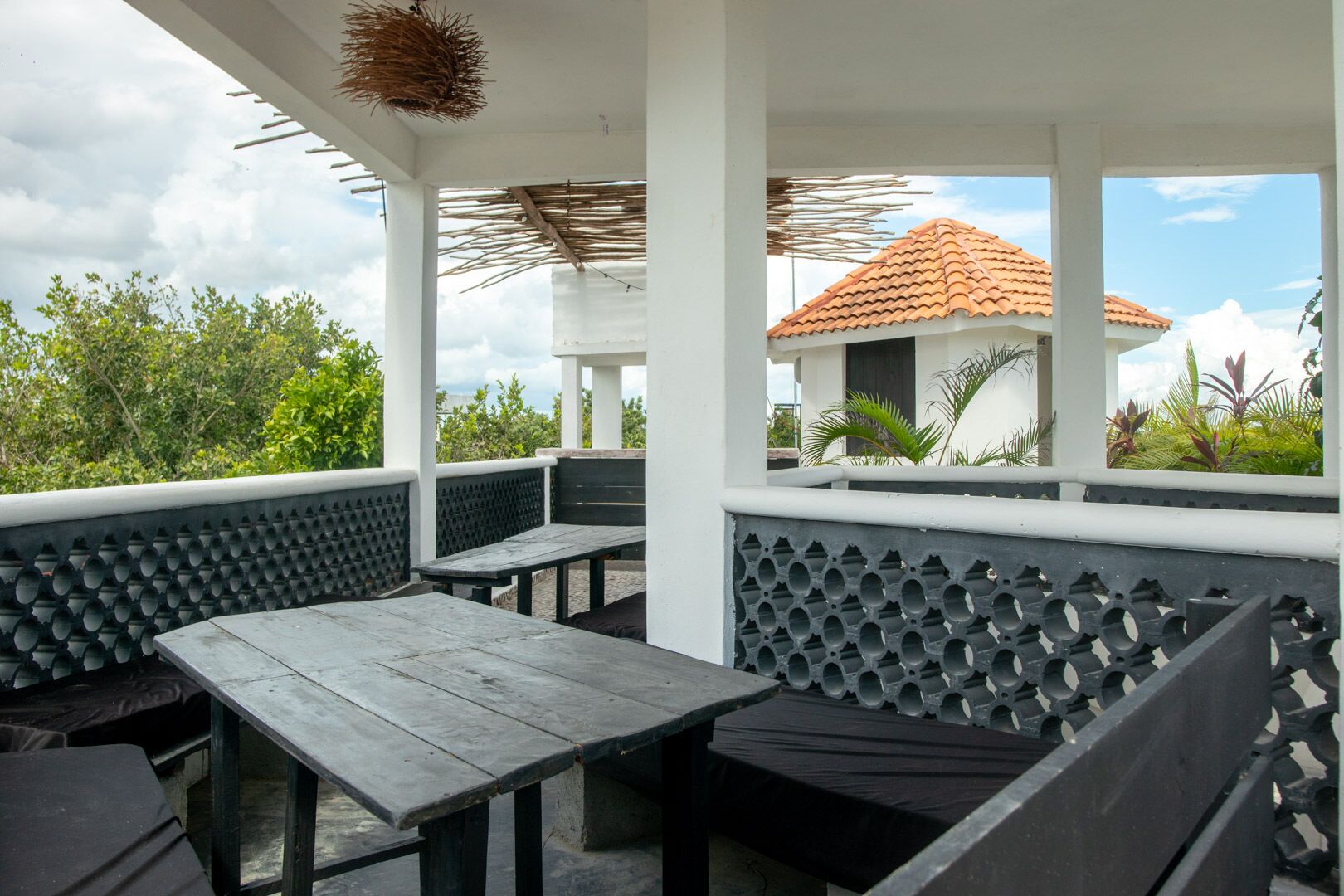 An outdoor seating area with wooden tables and benches, surrounded by white columns and a thatched roof.