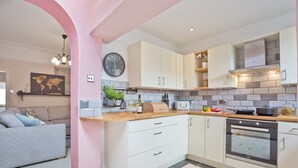 A modern kitchen with white cabinets, a built-in oven, and a dining area with a grey sofa. - Malleus House (Barrow-in-Furness)