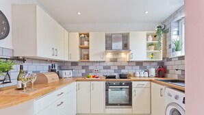 A modern kitchen with white cabinets, a built-in oven, a washing machine, and a variety of appliances and utensils on the countertops. - Malleus House (Barrow-in-Furness)