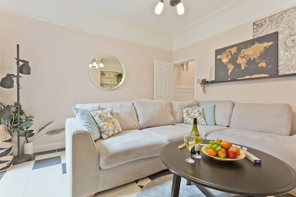 Een woonkamer met een beige bank, een ronde spiegel, een wereldkaart aan de muur en een tafel met fruit en wijnglazen.