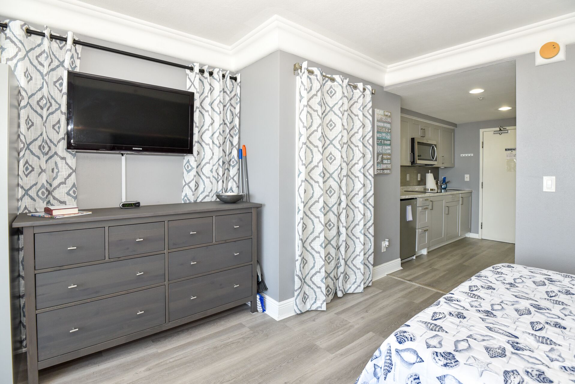 A bedroom with a TV, grey dresser, patterned curtains, and a bed with a blue and white design.