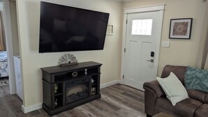 Een woonkamer met een flatscreen televisie aan de muur, een donker houten tv-meubel eronder, een bruine leren bank met een groene plaid, en een witte deur met een raam.
