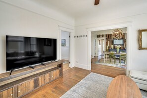 Un salon moderne avec un téléviseur à écran plat posé sur un meuble en bois, un coin repas avec une table et des chaises, et un couloir menant à une autre pièce.