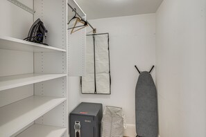 A storage closet with shelves, a hanging garment, a grey ironing board, and a black safe.