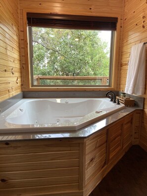 A wooden cabin bathroom with a large bathtub, a window with a view of trees, and a wooden deck outside.