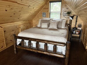 A cozy attic bedroom with a wooden sloped ceiling, a bed with white bedding, a nightstand with a lamp, and a heart-shaped decoration on the wall.