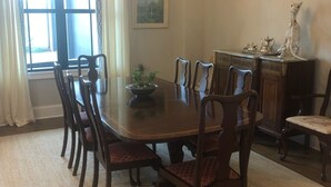 Uma sala de jantar com uma grande mesa de madeira, seis cadeiras, um lustre, uma janela com cortinas, um quadro na parede e um armário com objetos decorativos.
