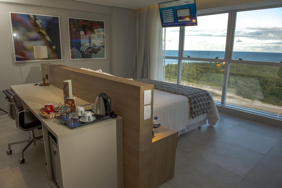 A hotel room with a desk, chair, bed, and large windows offering a view of the beach and ocean.