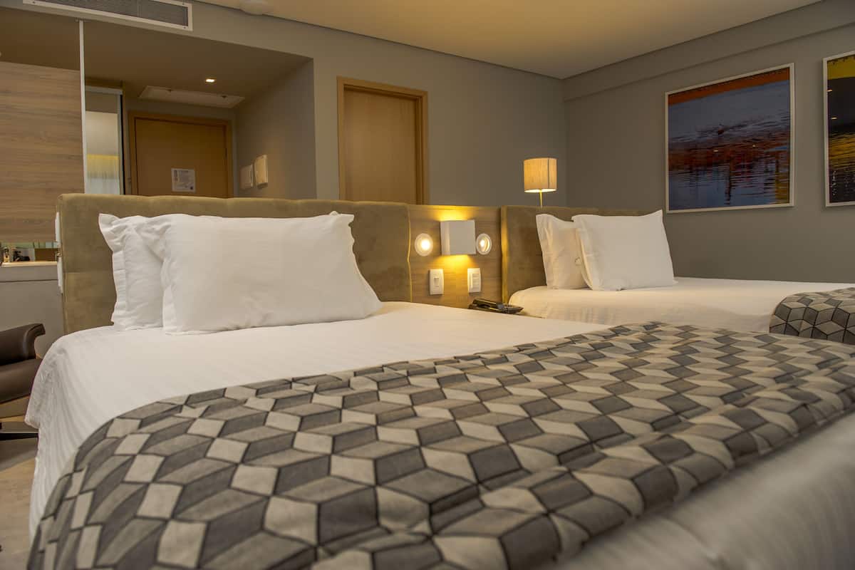 A hotel room with two beds, a large mirror, a bedside table with a lamp, and a painting on the wall.