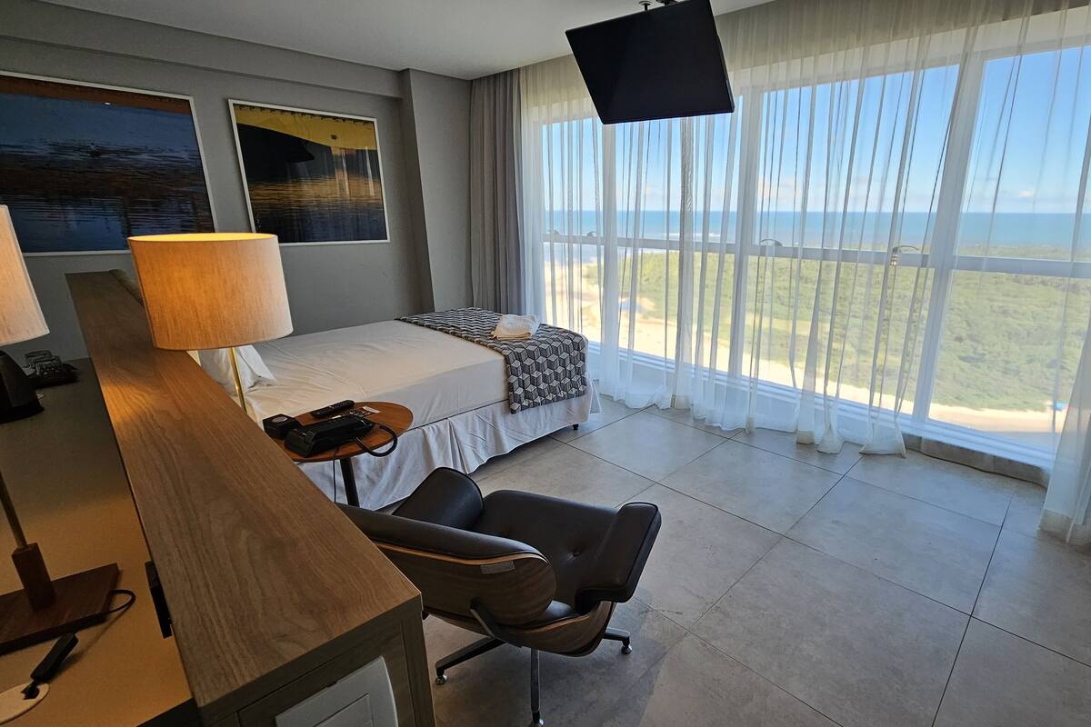 A hotel room with a bed, a desk with a lamp, a chair, a TV, and large windows with a view of the sea.