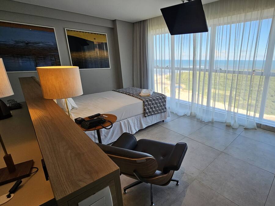 A hotel room with a bed, a desk with a lamp, a chair, a TV, and large windows with a view of the sea.