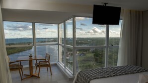 Hotelkamer met een groot raam met uitzicht op een waterpartij en groen. Er staat een bed, een aan de muur bevestigde televisie en een kleine tafel met stoelen.