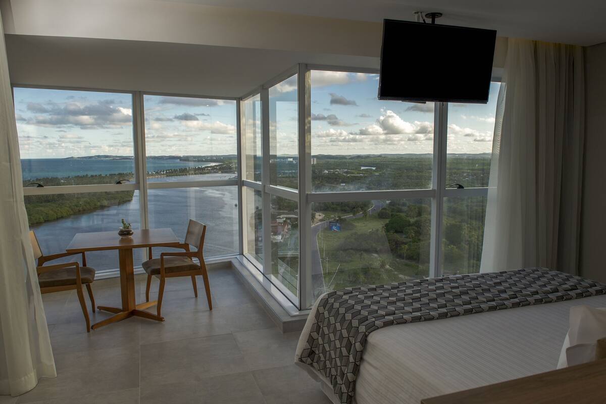A hotel room with a large window offering a view of a body of water and greenery. There is a bed, a TV mounted on the wall, and a small table with chairs.