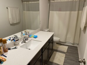 Une salle de bain avec un lavabo blanc, un miroir et des toilettes. On y trouve des articles de toilette sur le lavabo, un rideau de douche et un tapis sur le sol.