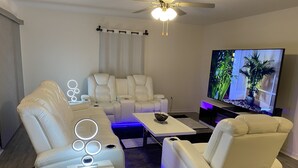 A modern living room with a flat-screen TV, white leather sofas, a coffee table, and a ceiling fan.