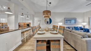 Une cuisine moderne avec des meubles blancs, un coin repas avec une table blanche dressée pour six personnes, et un canapé gris dans l’espace salon.