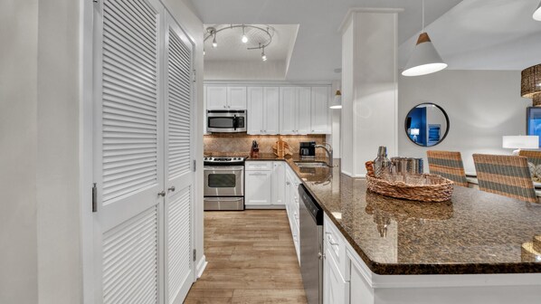A modern kitchen with white cabinetry, stainless steel appliances, a granite countertop, and a dining area with wicker chairs.