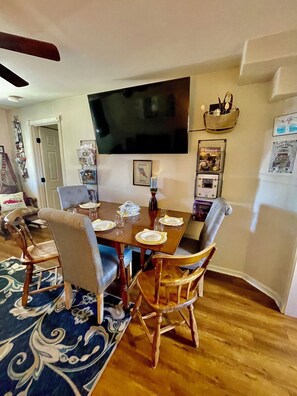 A dining area with a wooden table set for four, surrounded by upholstered chairs. A flat-screen TV is mounted on the wall above the table.