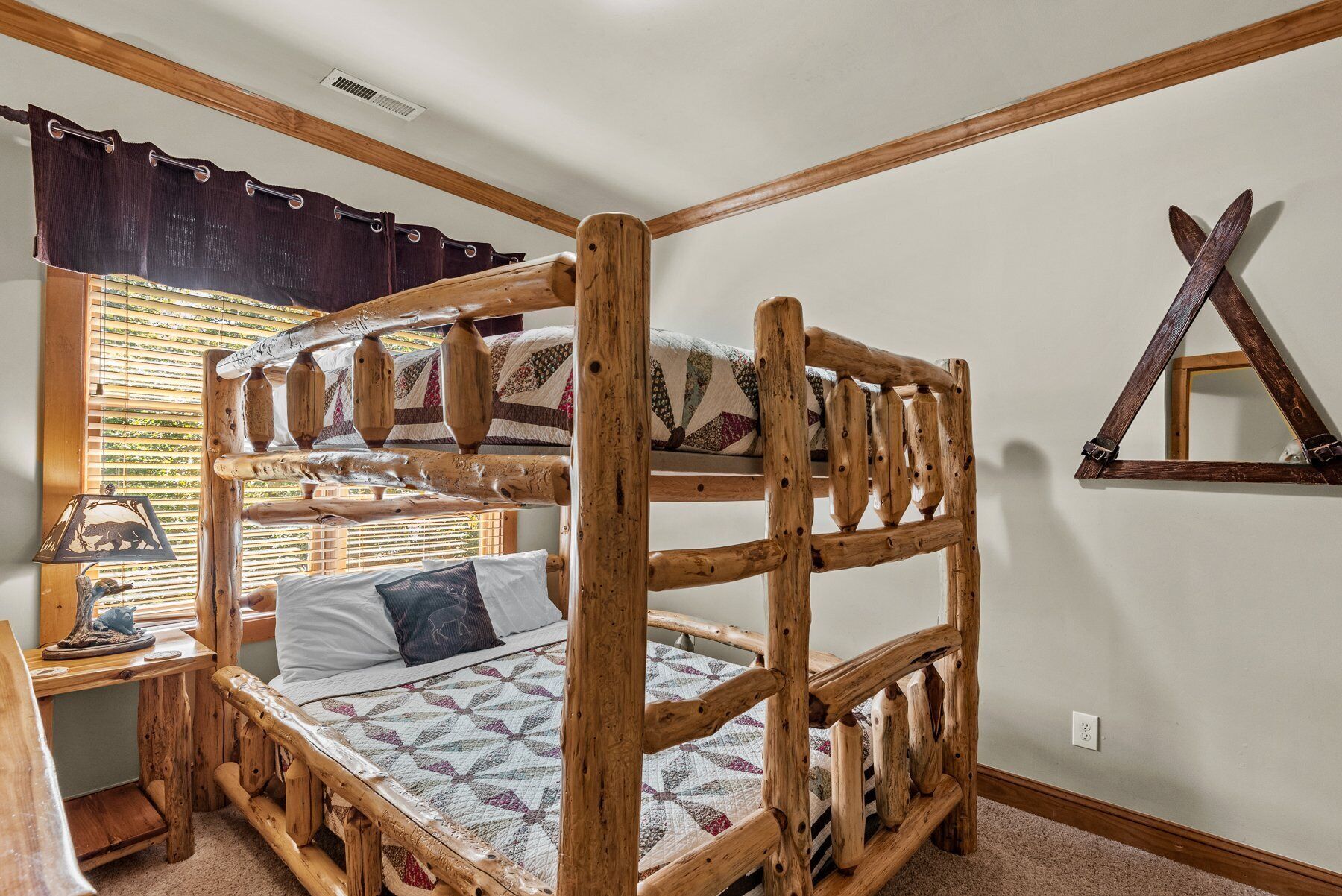 Un lit superposé avec une structure en bois et une couvre-lit matelassé. Il y a une fenêtre avec des stores, une lampe et une étagère fixée au mur, sur laquelle est posé un objet décoratif.