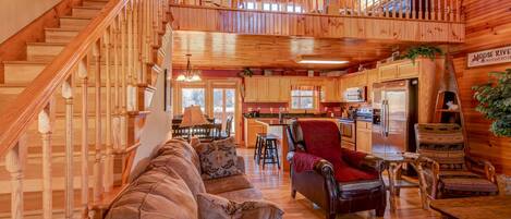 A cabin interior with a wooden staircase, a cozy living area with a sofa and armchairs, a kitchen with wooden cabinets, and a dining table.