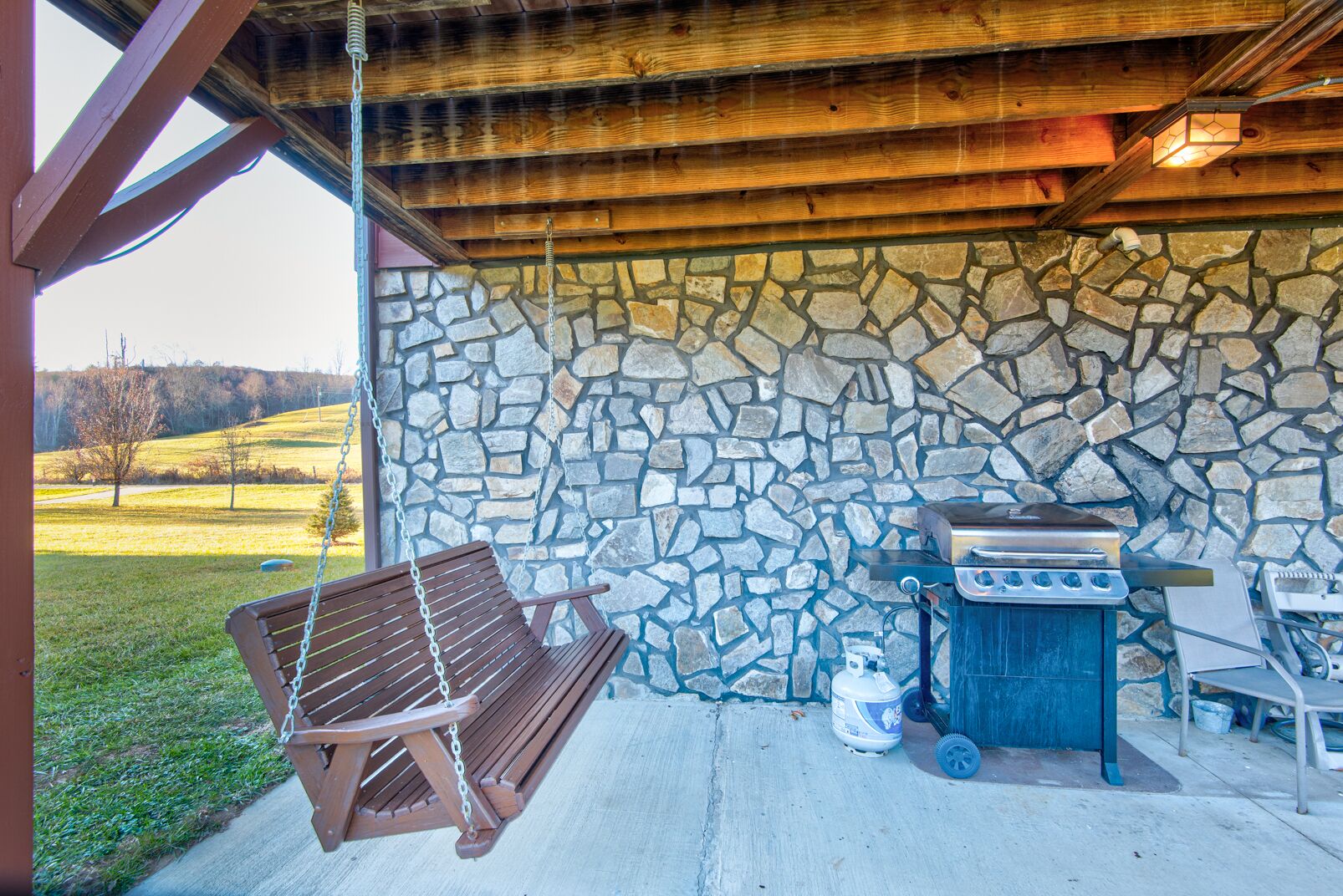 Un espacio al aire libre techado con pared de piedra, banca de madera colgante y una parrilla de barbacoa.