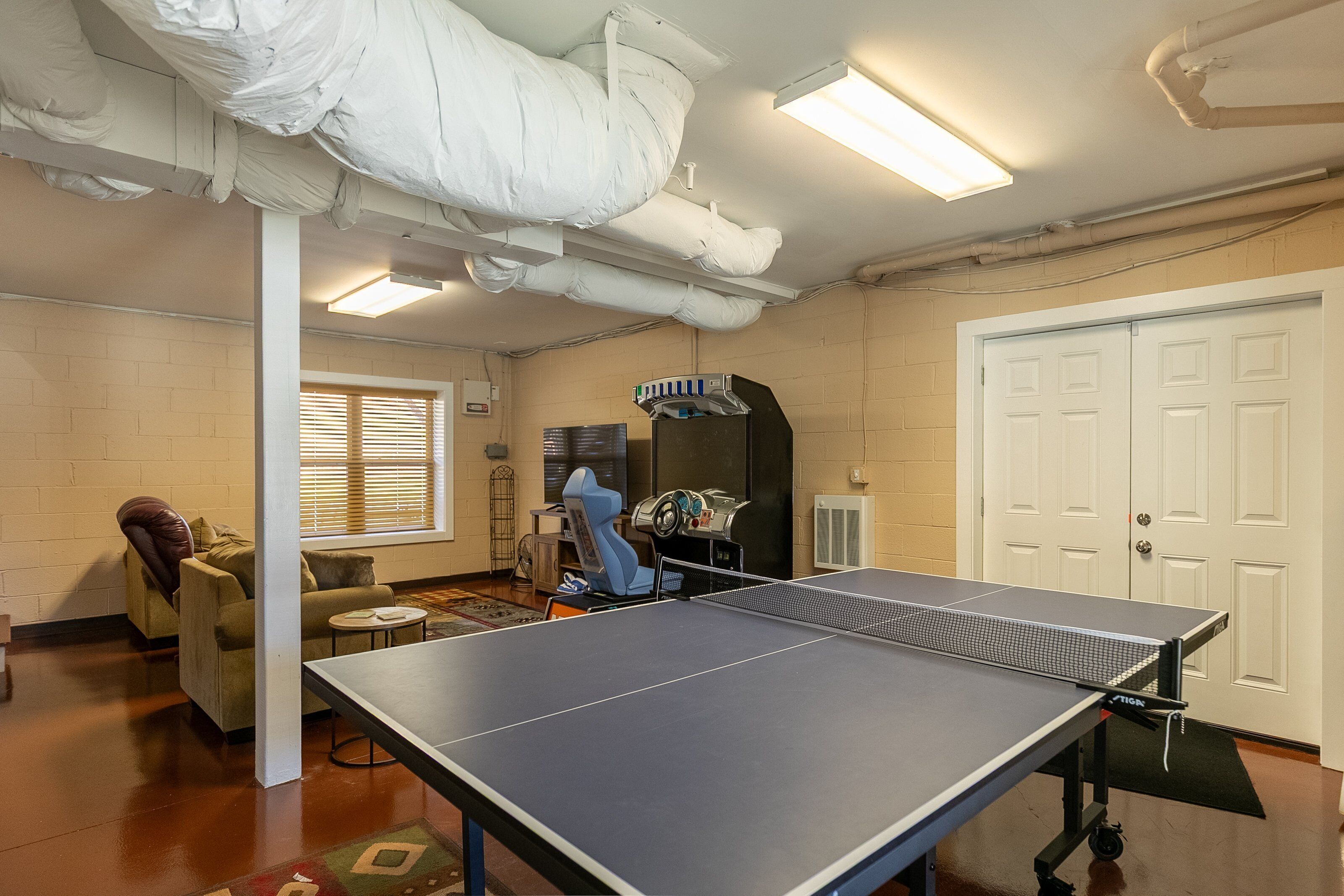 Una mesa de ping pong en una habitación con pared de ladrillo, una puerta y una ventana con persianas.