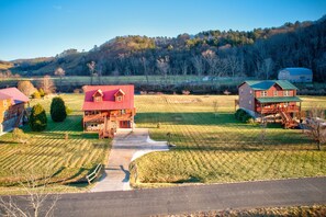 A large, two-story wooden house with a red roof and a balcony, surrounded by a well-maintained lawn and a forested hill in the background.