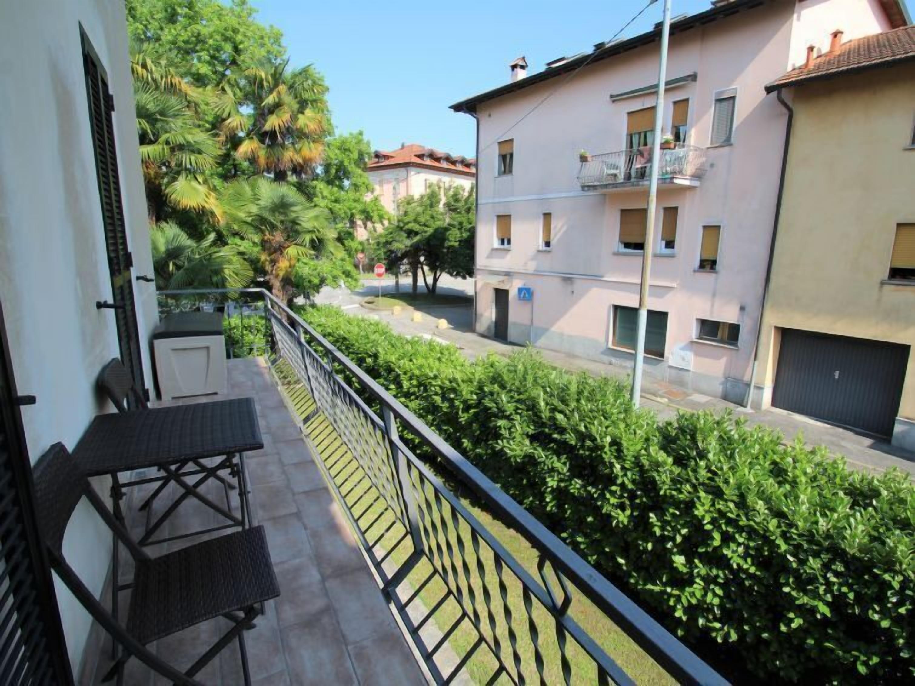 A balcony with a table and chairs, overlooking a street with buildings and greenery.