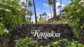 Eine steinerne Mauer mit der Aufschrift "Kanaloa" in weißer Schrift, umgeben von üppiger Vegetation und Palmen.
