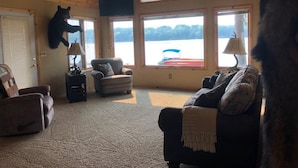 L’intérieur d’un chalet accueillant, avec une tête d’ours accrochée au mur, une cheminée, une grande fenêtre donnant sur un lac et un ventilateur de plafond.