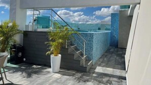 Un área de piscina moderna al aire libre con cerramiento de vidrio, una escalera que sube a un nivel superior y plantas en macetas.