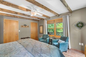 A bedroom with a bed, two chairs, a small table, a window with a view, and a ceiling fan.