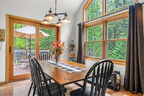 A dining area with a wooden table, black chairs, a vase with flowers, and large windows with curtains. - Dog-Friendly Three Little Birds Cottage on 3 Acres (Sturgeon Bay)